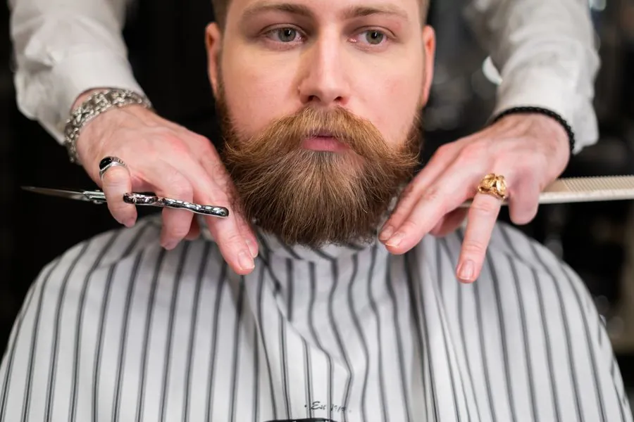 A bearded barber in a striped apron looking straight at the camera